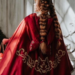 May include: A red hooded cape with a gold wolf design and gold embroidery. The cape has a white trim around the bottom edge.