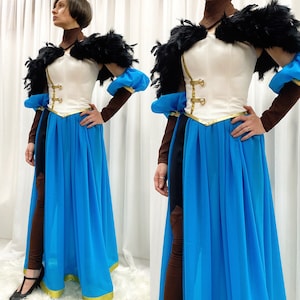 Pode incluir: Uma mulher vestindo um traje branco e azul com um colar de penas pretas. O traje apresenta detalhes dourados e uma longa saia azul.