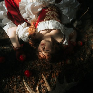 May include: A woman in a red and white dress lies on the forest floor surrounded by red apples. She has long red hair and is wearing a red corset with gold embroidery. The forest floor is covered in leaves and branches.