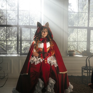 May include: A woman in a red hooded dress with a white lace trim and a fox ear headband. She is standing in a room with large windows and sunlight streaming in.