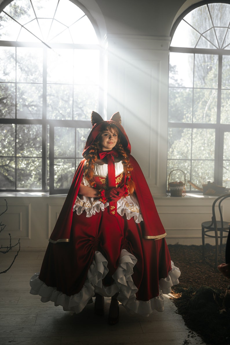 May include: A woman in a red hooded dress with a white lace trim and a fox ear headband. She is standing in a room with large windows and sunlight streaming in.