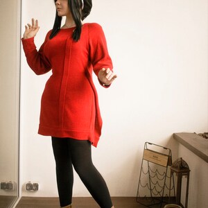 May include: A woman wearing a red sweater dress, black tights, and brown ankle boots. She is standing in a room with a white wall and a wooden floor.