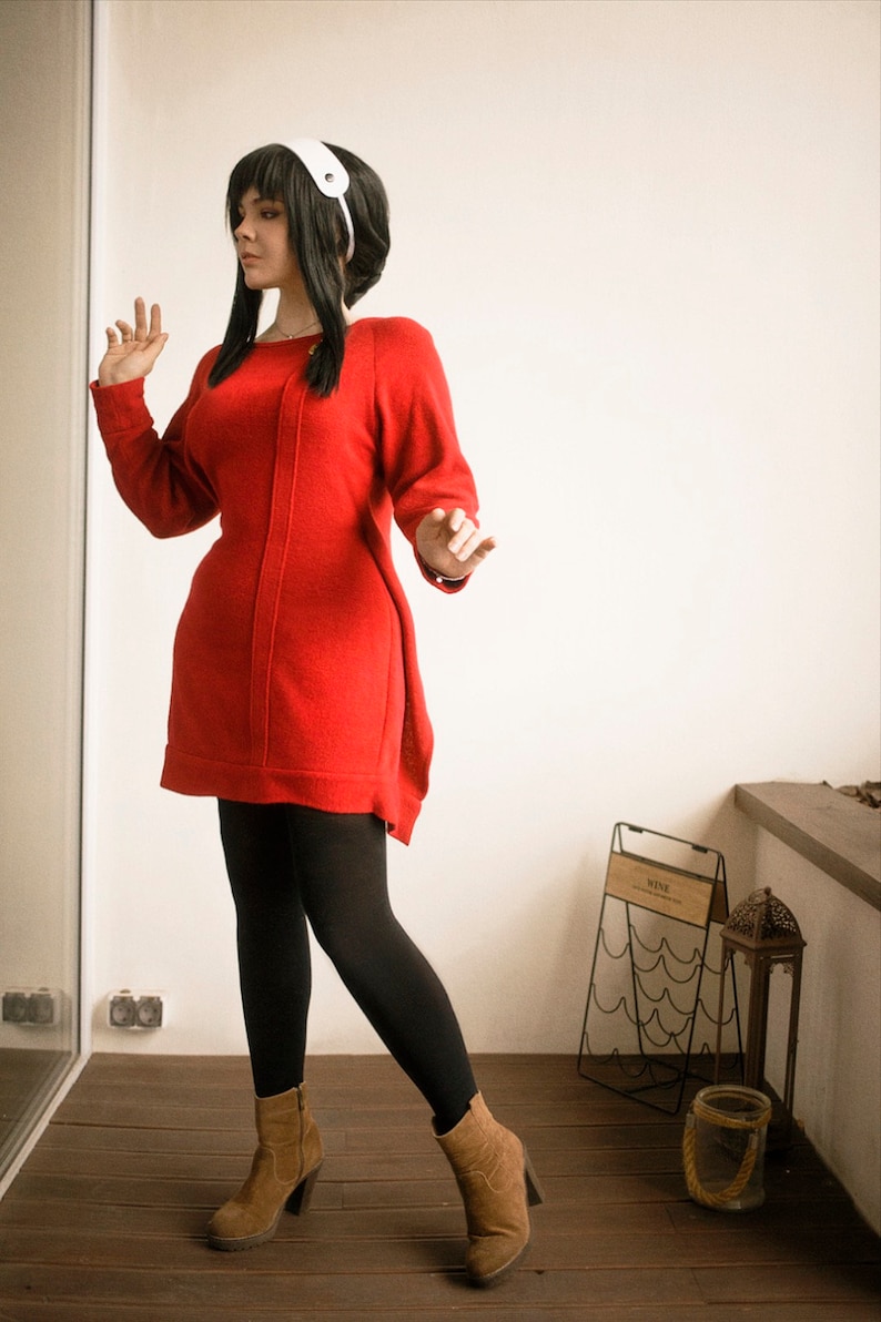 May include: A woman wearing a red sweater dress, black tights, and brown ankle boots. She is standing in a room with a white wall and a wooden floor.