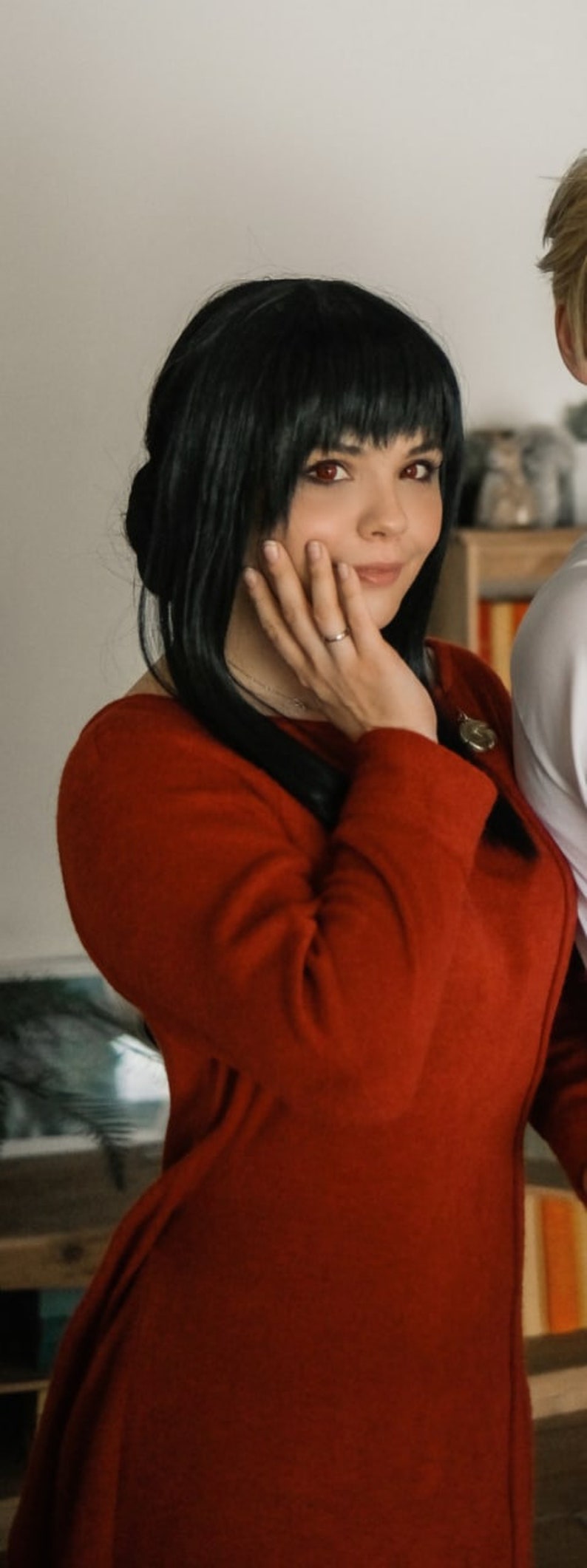 May include: A woman with long black hair wearing a red sweater. She is smiling and has her hand on her cheek.