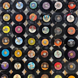 May include: A collection of vintage vinyl records in various colors and designs. The records are arranged in a grid pattern on a textured surface. Some of the records have text on the labels, including "Atlantic", "Arista", and "AM".
