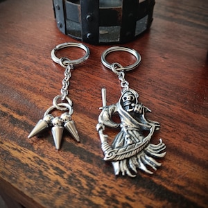 May include: Two silver-toned metal keychains. One keychain features a dangling charm with three pointed spikes attached to a circular ring. The second keychain features a detailed charm of a hooded skeleton figure holding a scythe.