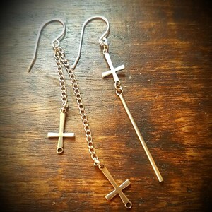 May include: Gold-coloured earrings with inverted cross pendants. One earring has a chain with two crosses, the other a single cross on a straight bar. The earrings hang from silver-coloured ear wires. The background is a dark wooden surface.