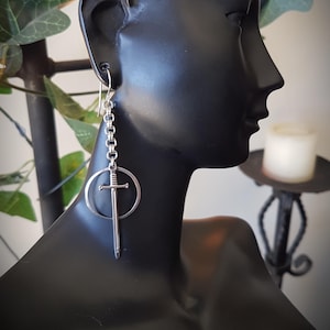 May include: Silver earrings with a sword pendant inside a circle, hanging from a chain. The earrings are shown on a black mannequin, highlighting their design. The sword has a detailed hilt and blade. The earrings are a unique accessory.
