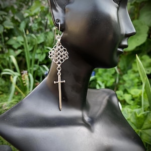 May include: A silver-toned earring featuring a chainmaille square component connected to a small sword charm. The earring hangs from a standard hook wire, showcasing a metallic finish and intricate interlocking ring patterns above the sword.