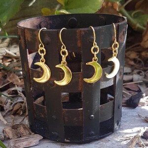 May include: Four gold-coloured crescent moon earrings displayed inside a dark, rustic metal container. The earrings feature a simple design with a hook and chain. The moons have a gold finish, reflecting light. The container has a square shape.