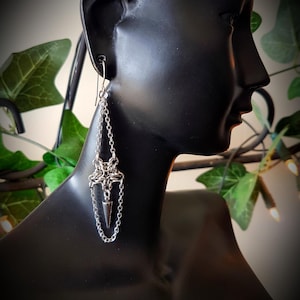 May include: Silver-coloured dangle earrings with a chain design. The earrings feature a star-like top, a chain, and a small, pointed pendant. The earrings are displayed on a black mannequin head, with green foliage in the background.