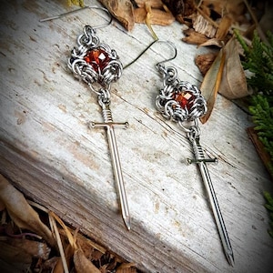 May include: A pair of silver earrings with a medieval design. Each earring has a red gemstone set in a woven metal design, with a small sword pendant hanging below. The earrings are presented on a wooden surface.