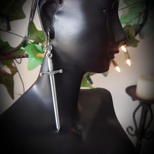 May include: A pair of silver-coloured sword earrings on a black mannequin. The earrings have a detailed sword design with a long, slender blade and a decorative hilt. They hang from a simple hook and ring closure. The background features greenery and soft lighting.