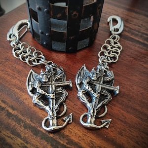 Peut inclure: Deux porte-clés en métal argenté représentant une figurine de gargouille détaillée agrippant une croix qui se prolonge en forme d'ancre. Chaque breloque est fixée à un segment de chaîne en argent et à un fermoir mousqueton en métal.