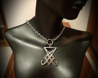 Può includere: Un ciondolo in metallo color argento raffigurante il Sigillo di Lucifero, sospeso a una catenina abbinata color argento. Il ciondolo presenta un design geometrico con una parte superiore triangolare e una base angolare simmetrica su un anello.