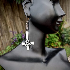 May include: A silver-toned earring featuring a hoop connected to a chain link segment that suspends an inverted cross pendant. The centre of the cross contains a circular cutout with a five-pointed star pentagram design, all in a polished metal finish.