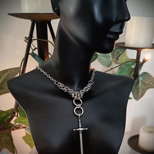 May include: A silver necklace with a sword pendant is shown on a black mannequin. The necklace has a chainmail-style collar, three connected rings, and a detailed sword pendant. The background includes candles and greenery.