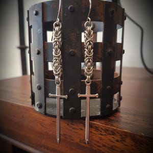May include: Silver earrings with a chainmail design and sword pendants. The earrings hang from hooks and are set against a dark, textured backdrop. The swords have a cross-shaped hilt and a pointed blade. The earrings are approximately 7 cm in length.