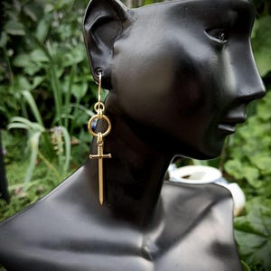Peut inclure: Une boucle d'oreille dorée composée d'un pendentif en forme d'épée suspendu à un anneau circulaire et un crochet. Le charm épée présente une garde détaillée et une lame droite, pendant verticalement au lobe d'un mannequin noir.
