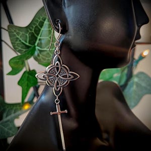 May include: Silver Celtic knot earrings with a dangling sword charm. The earrings feature intricate knotwork and a detailed sword pendant. The earrings are displayed on a black mannequin head, with green foliage in the background.