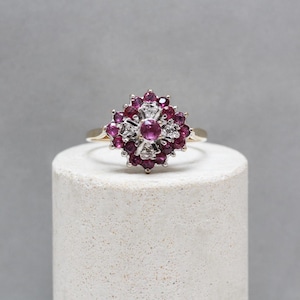 Könnte beinhalten: Ein goldfarbener Ring mit einem floralen Muster aus roten Edelsteinen und kleinen weißen Steinen. Der Ring ist auf einem weißen zylindrischen Ständer vor grauem Hintergrund platziert.