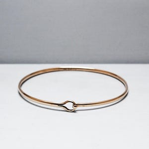 May include: A delicate gold-colored bangle bracelet. The bracelet has a simple, thin design with a hook-and-eye clasp. The jewelry is displayed on a white surface with a soft, neutral background.