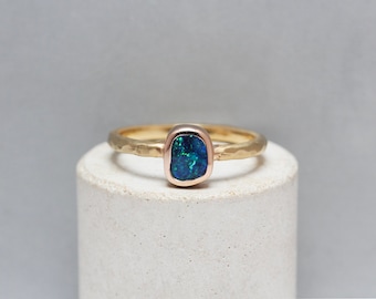 May include: A gold ring with a blue-green opal gemstone set in a bezel setting. The ring band is hammered for a textured look.