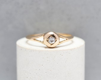Könnte beinhalten: Ein goldener Ring mit einem einzelnen runden Diamanten in einer Zargenfassung. Der Ring hat ein schlichtes, elegantes Design.