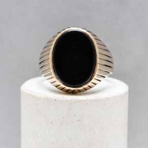 May include: A silver ring with a black oval gemstone set in the center. The band of the ring has a ridged design.