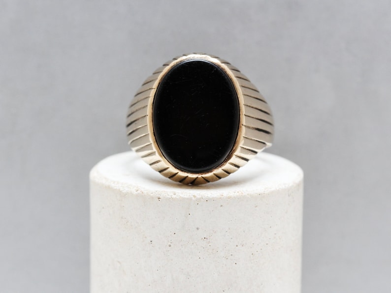May include: A silver ring with a black oval gemstone set in the center. The band of the ring has a ridged design.