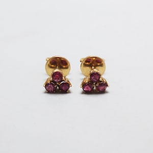 May include: A pair of gold-colored stud earrings, each featuring three small, round, deep pink gemstones arranged in a triangular cluster. The earrings are set against a plain white background.