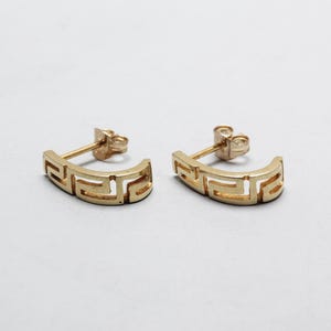 May include: A pair of gold-colored stud earrings. Each earring features a curved design with a repeating geometric pattern. The earrings are displayed on a white background.