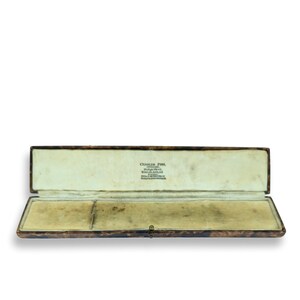 Puede incluir: Una caja de joyería de cuero marrón abierta y vacía con un interior de terciopelo blanco. La caja tiene el texto "Charles Fish, Goldsmith, 15, Old Bond Street, London, W.1. Telephone: HYDe Park 2274" impreso en el interior.