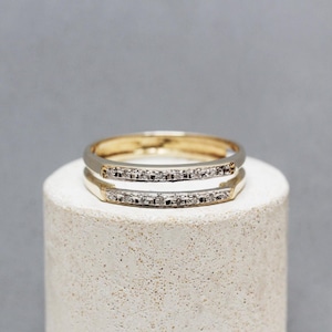 Könnte beinhalten: Zwei goldene Ringe mit einer Reihe kleiner, klarer Steine. Die Ringe sind übereinander gestapelt und ruhen auf einem weißen, zylindrischen Sockel vor grauem Hintergrund. Schmuck.