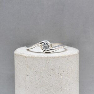 Puede incluir: Un anillo de plata con una gema redonda y transparente engastada en un diseño en espiral. El anillo se muestra sobre un soporte cilíndrico blanco sobre un fondo gris. La gema es el punto focal del anillo.
