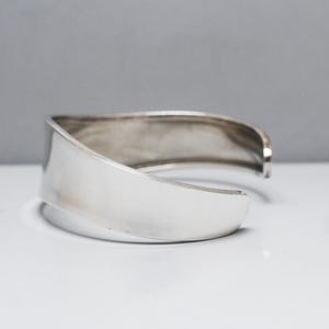 May include: A wide, polished silver cuff bracelet with a smooth, reflective surface and a curved, open-ended design. The metal band features a consistent width and a slightly undulating edge, showcasing a clean, metallic finish throughout the piece.