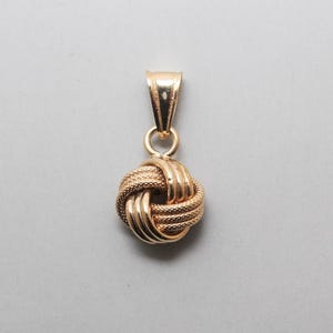 May include: A gold pendant featuring a spherical knot design. The pendant is composed of alternating smooth and textured gold bands woven together into a circular shape, suspended from a tapered, polished gold bail with a small jump ring.