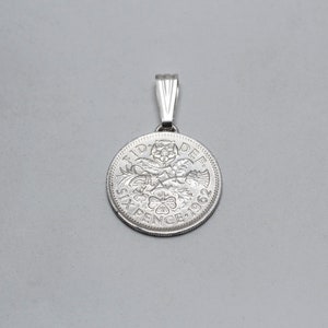 Puede incluir: Colgante de moneda de seis peniques de plata con la inscripción "FID DEF SIXPENCE 1962" y un diseño de una figura coronada que sostiene un cetro y un escudo.