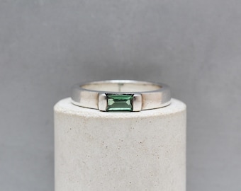 May include: A silver ring featuring a rectangular, emerald-green gemstone. The ring is displayed on a white cylindrical stand against a grey backdrop. The gemstone is the focal point, displaying a vibrant green hue.