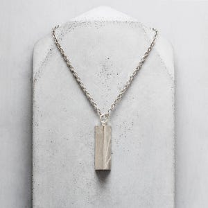 Può includere: Una collana color argento con un pendente rettangolare. Il pendente è di colore grigio chiaro e pende da una catena. La collana è esposta su una superficie bianca strutturata.