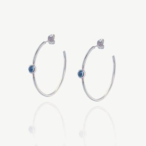Peut inclure: Une paire de boucles d'oreilles créoles en argent, chacune ornée d'une petite pierre précieuse bleue et ronde. Les boucles d'oreilles ont un design simple et élégant, avec une finition polie. Les pierres sont serties au centre des créoles.