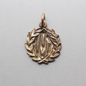 May include: A circular gold-toned metal pendant featuring a raised laurel wreath border surrounding a central design of three vertical, elongated, pointed shapes. The pendant includes a small circular jump ring at the top for attachment.