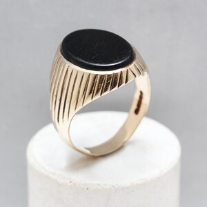 May include: A gold ring with a black oval gemstone set in the center. The band of the ring has a ridged design.