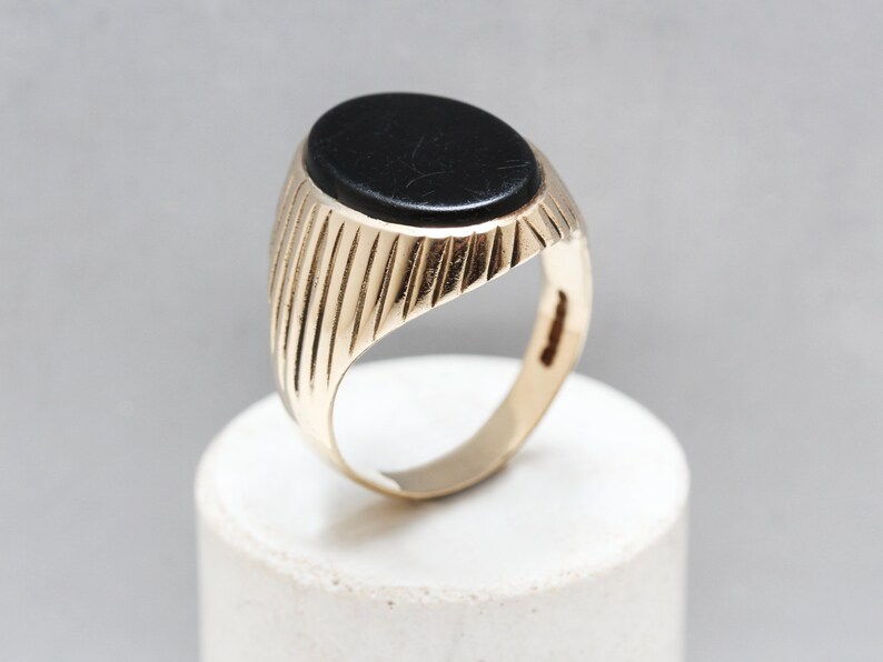 May include: A gold ring with a black oval gemstone set in the center. The band of the ring has a ridged design.
