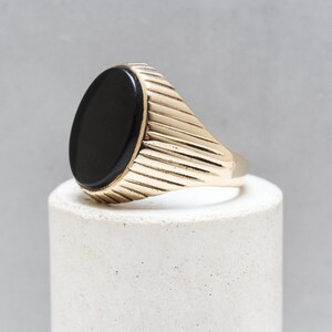 May include: A gold ring with a black oval gemstone. The band of the ring has a ridged design.