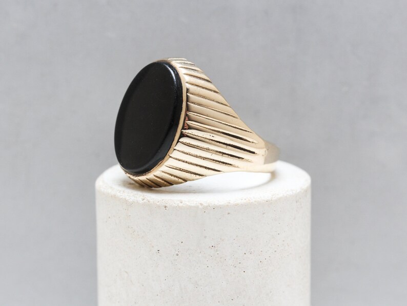 May include: A gold ring with a black oval gemstone. The band of the ring has a ridged design.