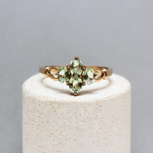 Könnte beinhalten: Ein goldfarbener Ring mit fünf hellgrünen, oval geschliffenen Edelsteinen. Das Ringband hat ein filigranes, verschlungenes Design. Die Edelsteine sind in einem floralen Muster angeordnet, was einen klassischen und eleganten Look erzeugt.