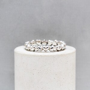 Peut inclure: Une bague en argent ornée d'un motif de cœurs répétés. La bague est présentée sur un support cylindrique blanc, sur fond gris. Le motif en forme de cœur de la bague est visible en gros plan.