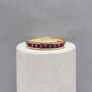 May include: A gold band ring with a row of square-cut, deep pink gemstones. The ring is set on a white cylindrical pedestal against a gray background. The ring's design is simple and elegant.