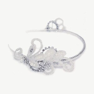 Puede incluir: Una diadema de novia plateada con un diseño floral. La diadema presenta una delicada banda curva adornada con cristales brillantes y perlas de imitación. Los adornos florales están intrincadamente detallados.